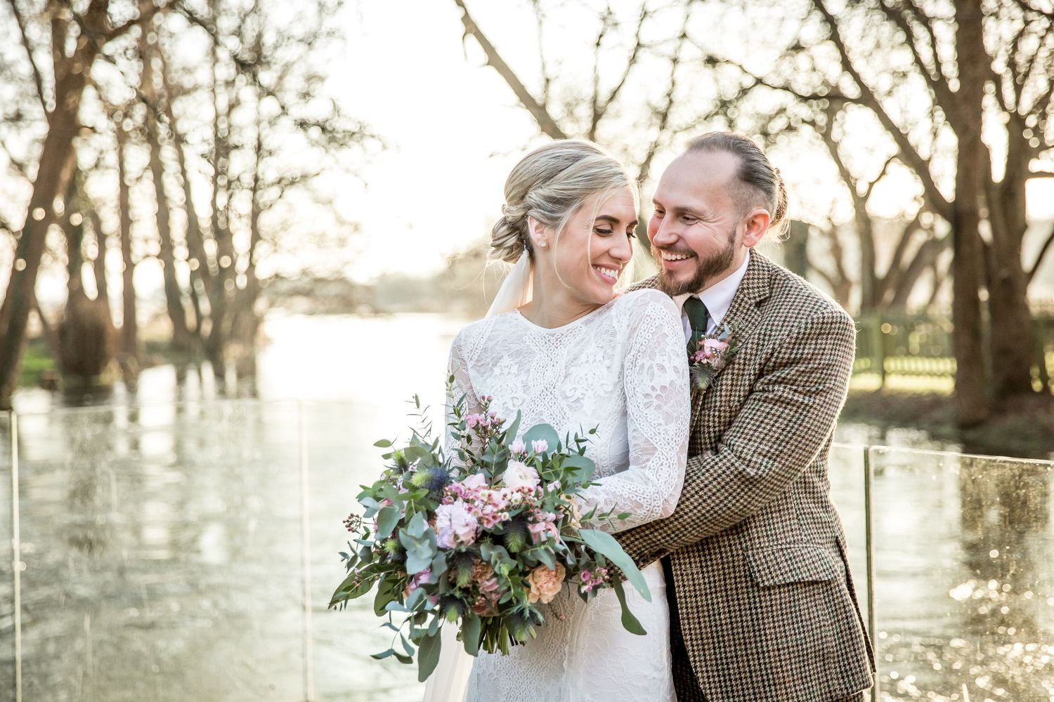 A couple in wedding attire embraces outdoors with a lush bouquet of flowers near a waterfront at sunset.
