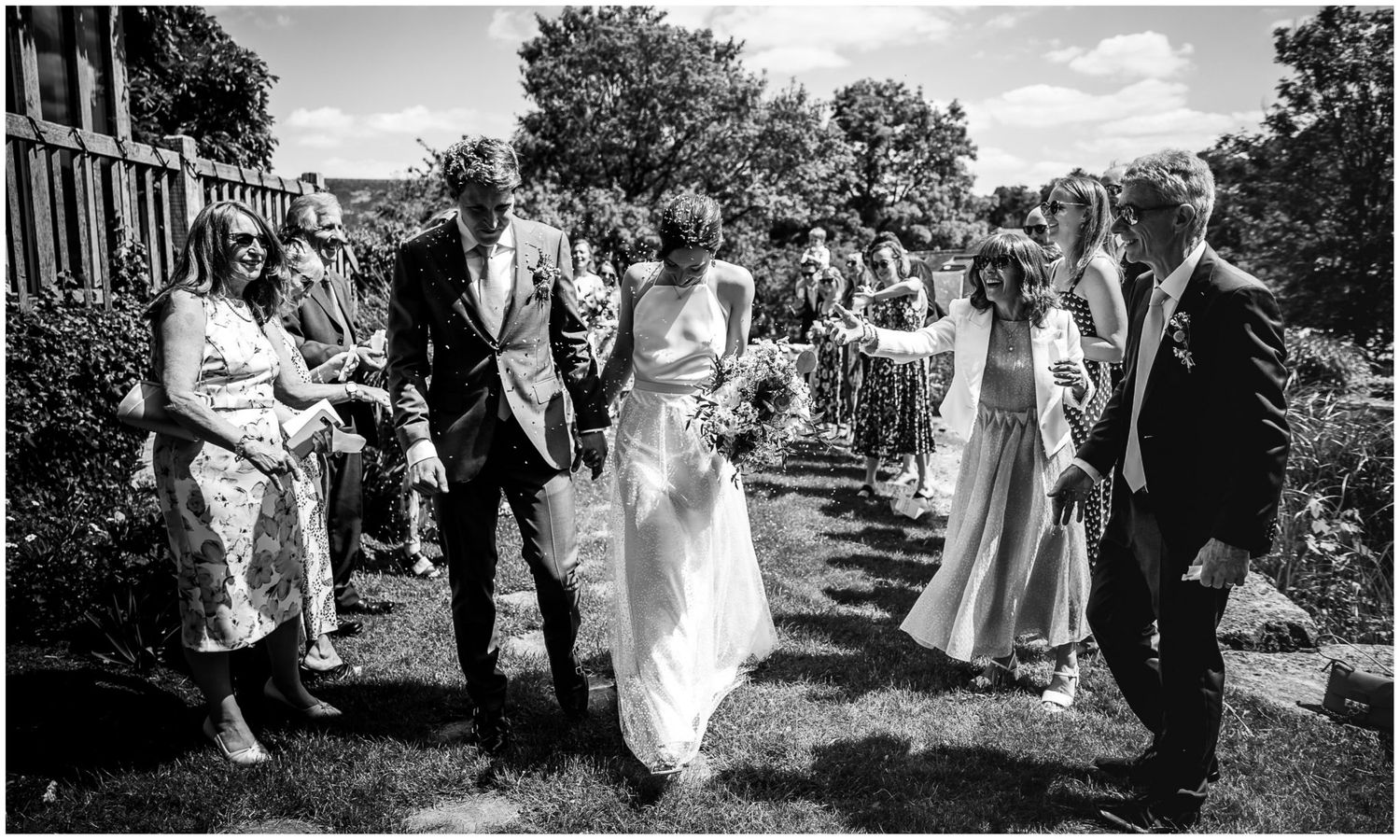 Wedding party and guests gather for outdoor celebration in garden setting.