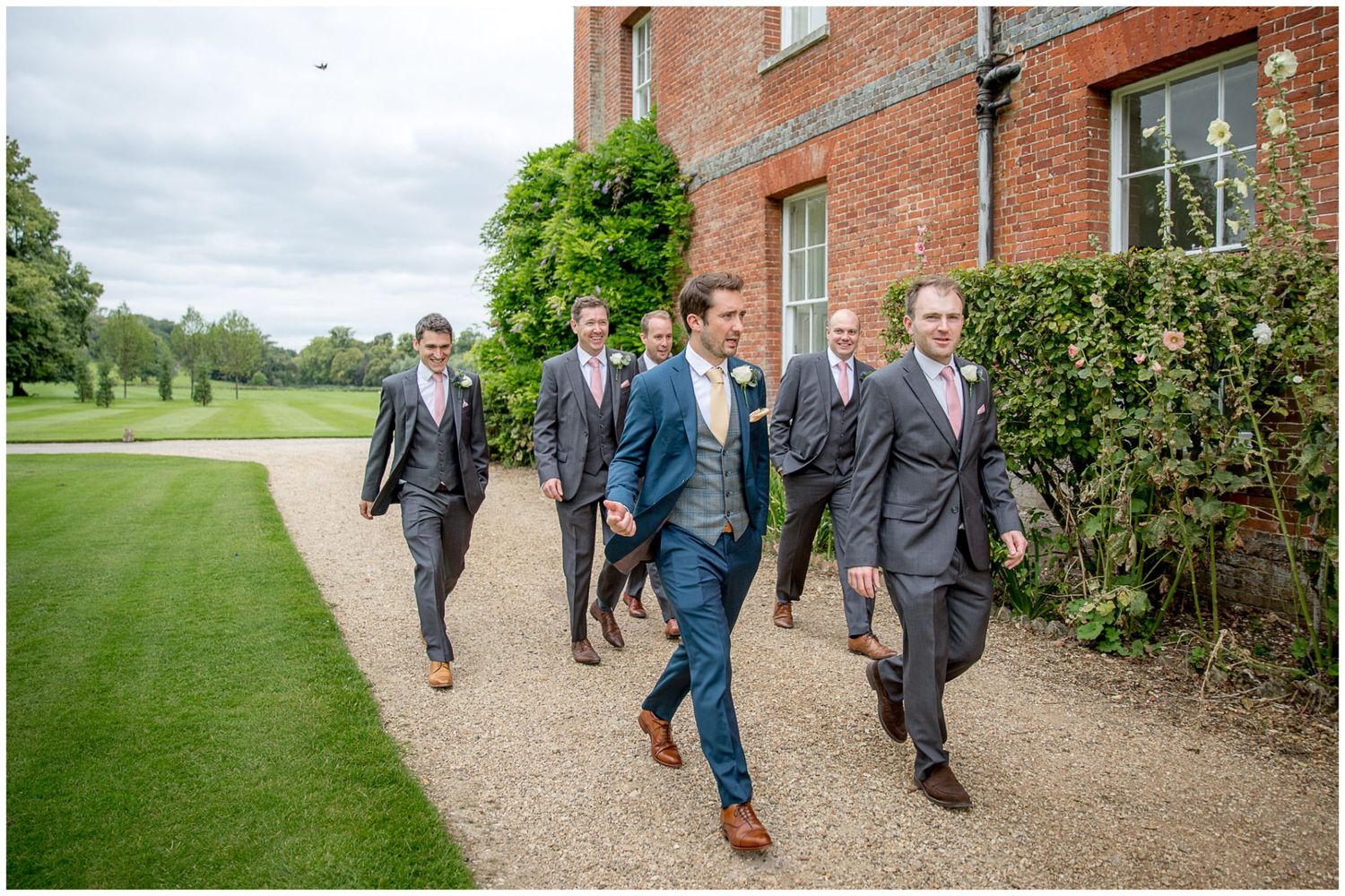 Wedding party walks along brick building pathway surrounded by green landscaping.
