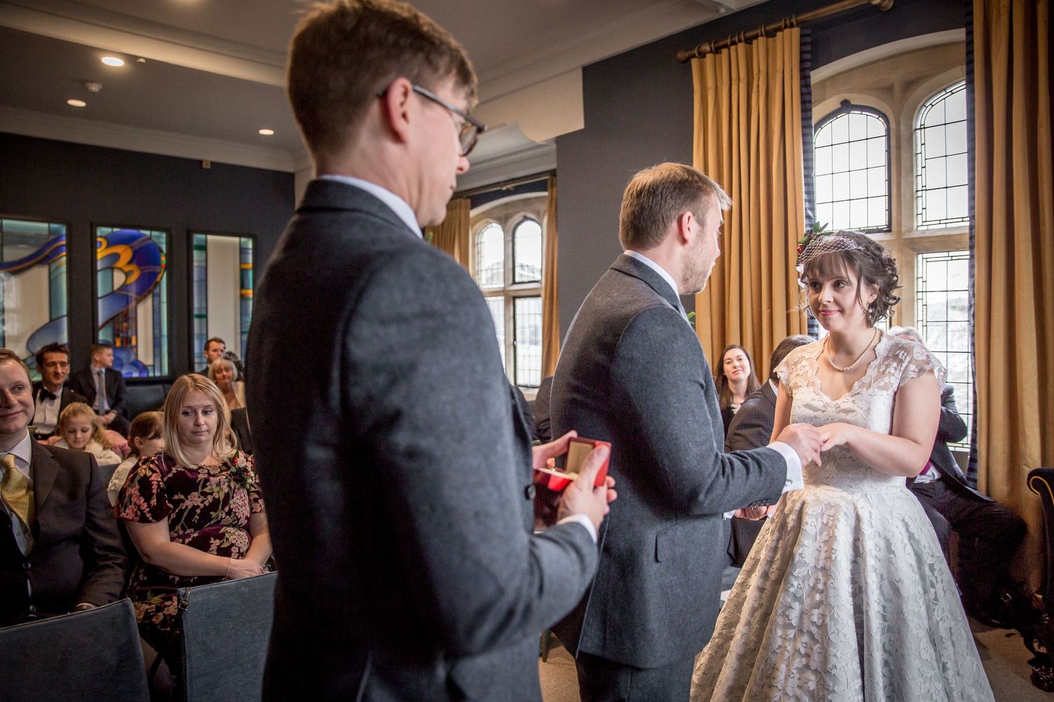 Wedding ceremony in progress inside historic building with arched windows and gathered guests.