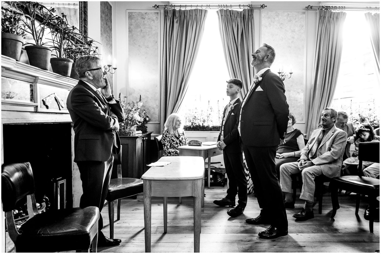 Black and white photo of ceremony proceedings in an elegant room with curtains.