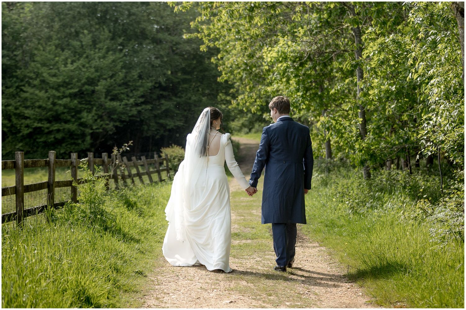 Newlyweds walking hand in hand down a country path beside a wooden fence.