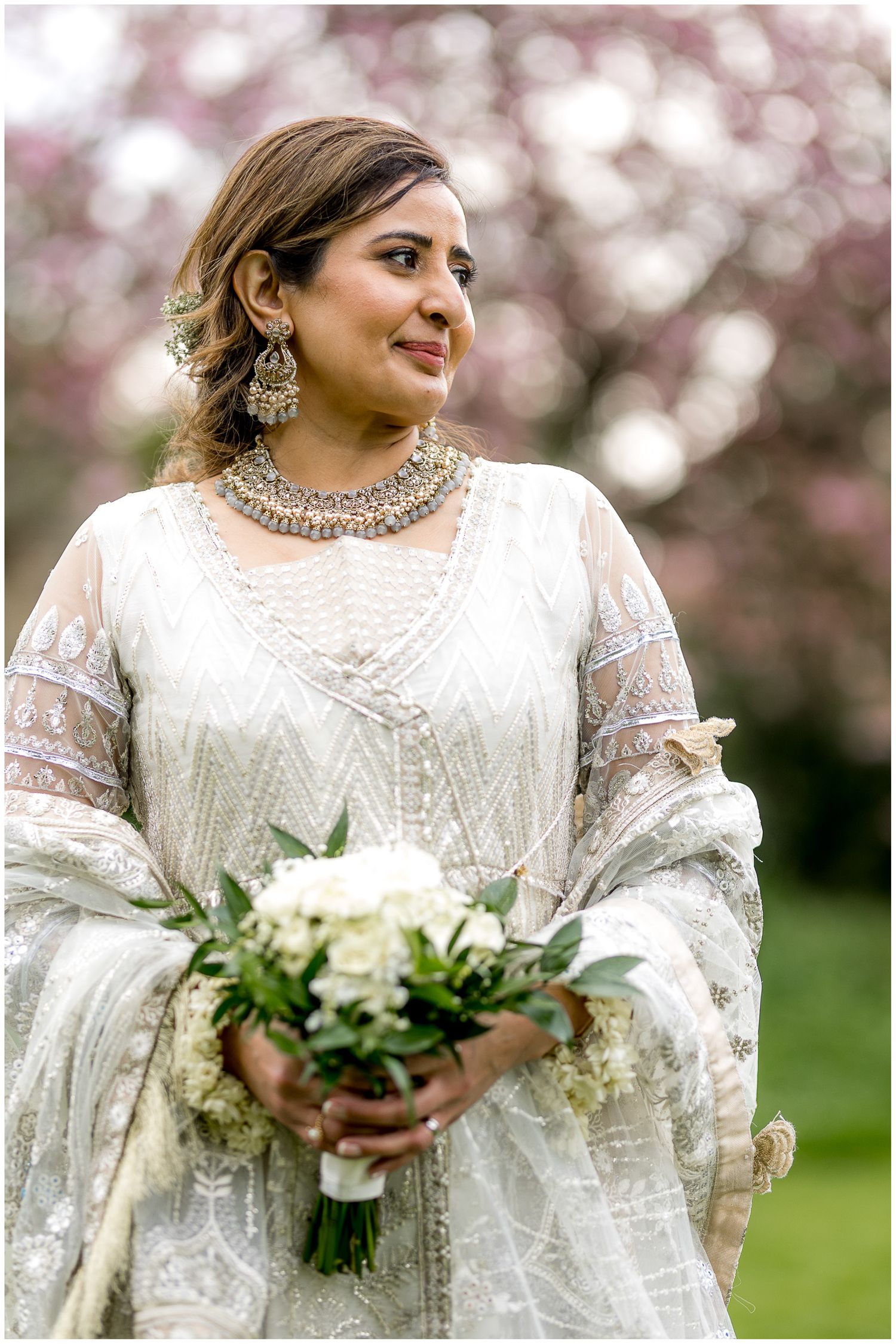 Close-up details of a white lace wedding dress and bouquet against a soft pink floral background.