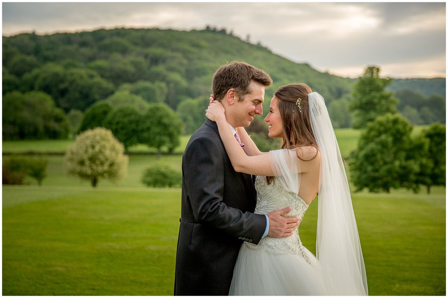 A romantic wedding couple embraces on a lush green hillside during sunset.