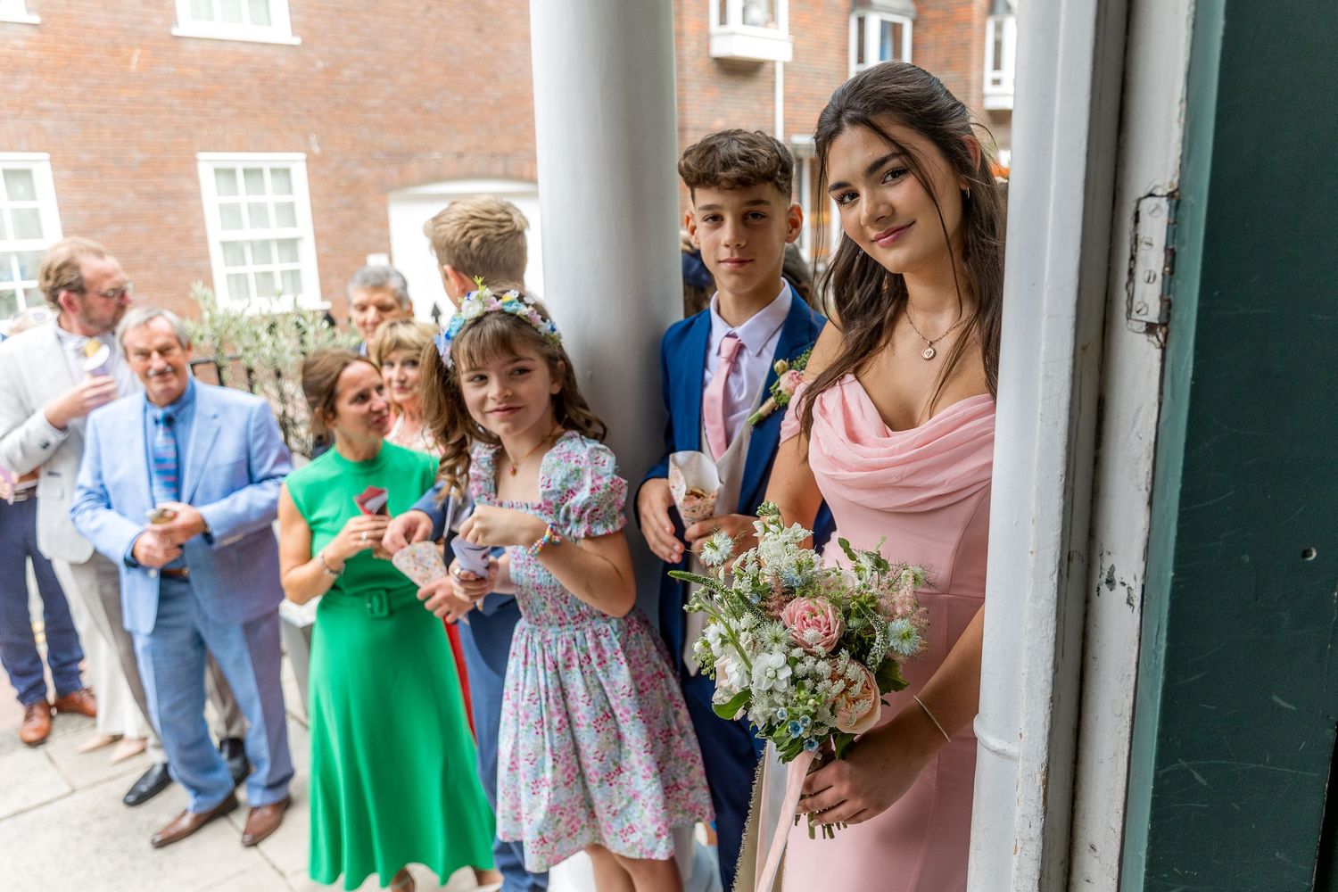Wedding party and guests gathered under white columns outside brick building.
