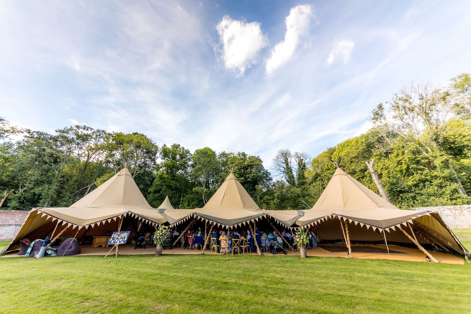 Panoramic view of multiple tipi-style wedding tents set up in a green field under blue skies.