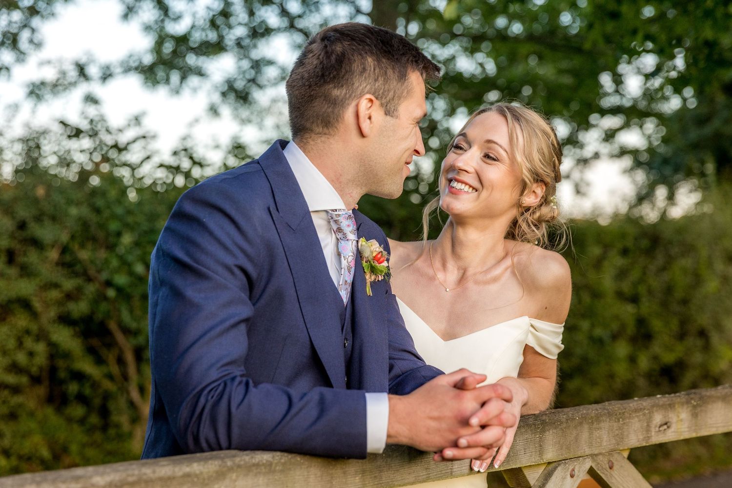 A couple laughs together while leaning against a wooden fence in a garden setting.