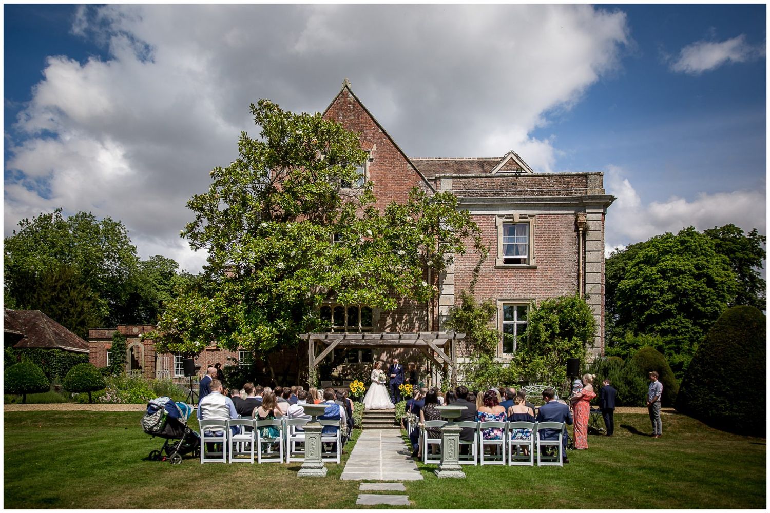 Outdoor wedding ceremony setup with white chairs and guests gathered on a beautiful lawn in front of a historic brick building.