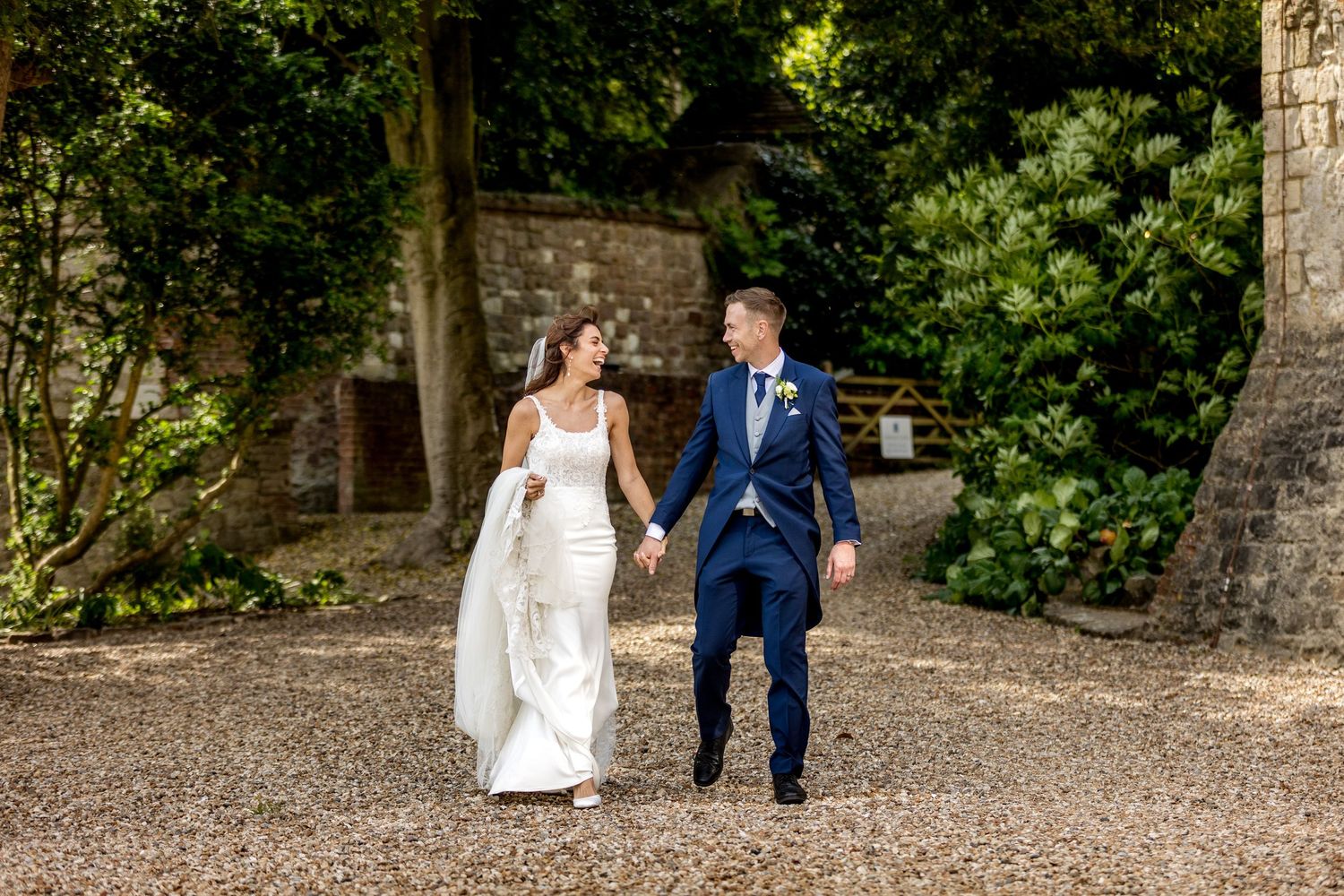 Bride in white dress and groom in navy suit walking through garden path during wedding portraits.