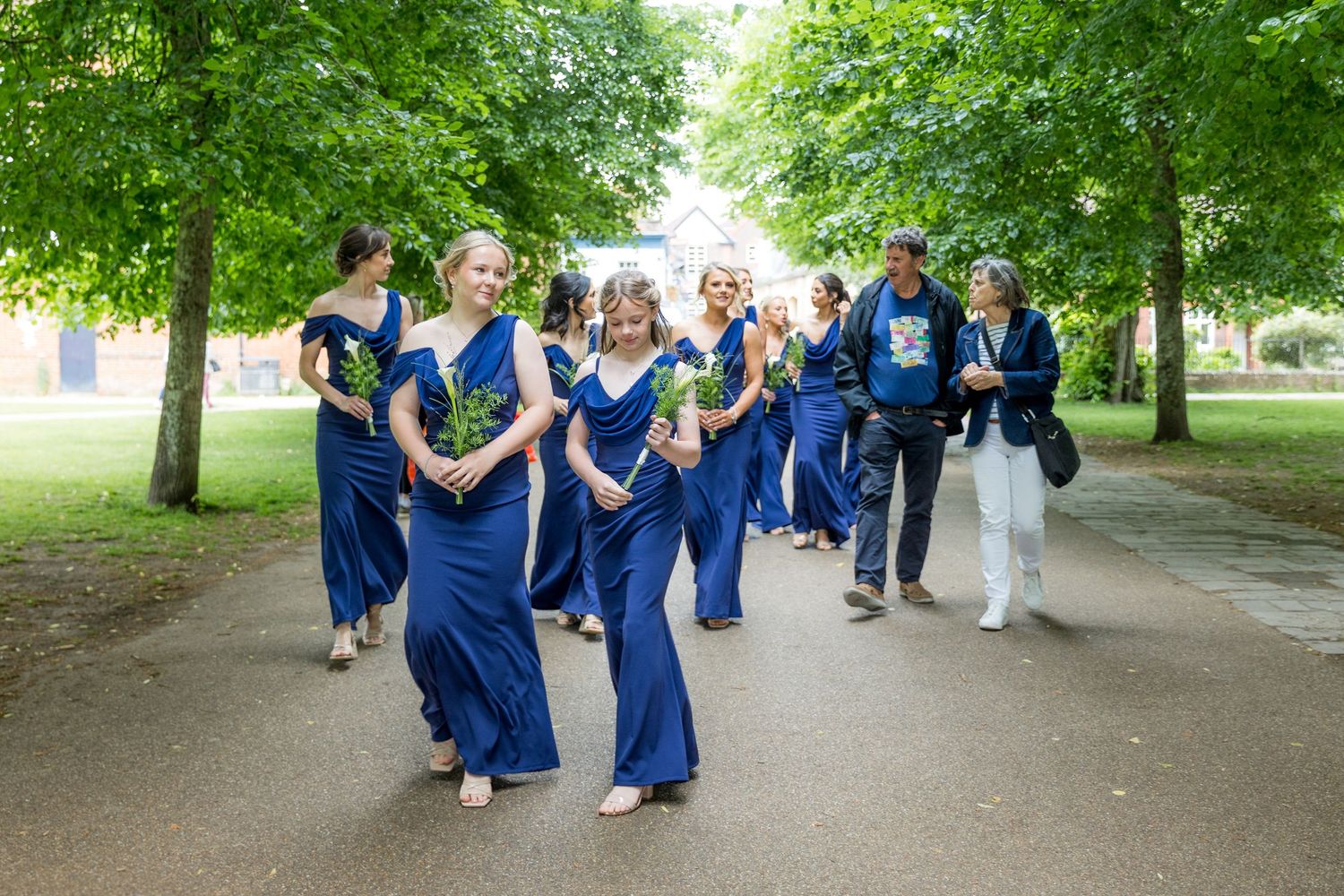 Wedding party in navy blue attire walking through a tree-lined path.