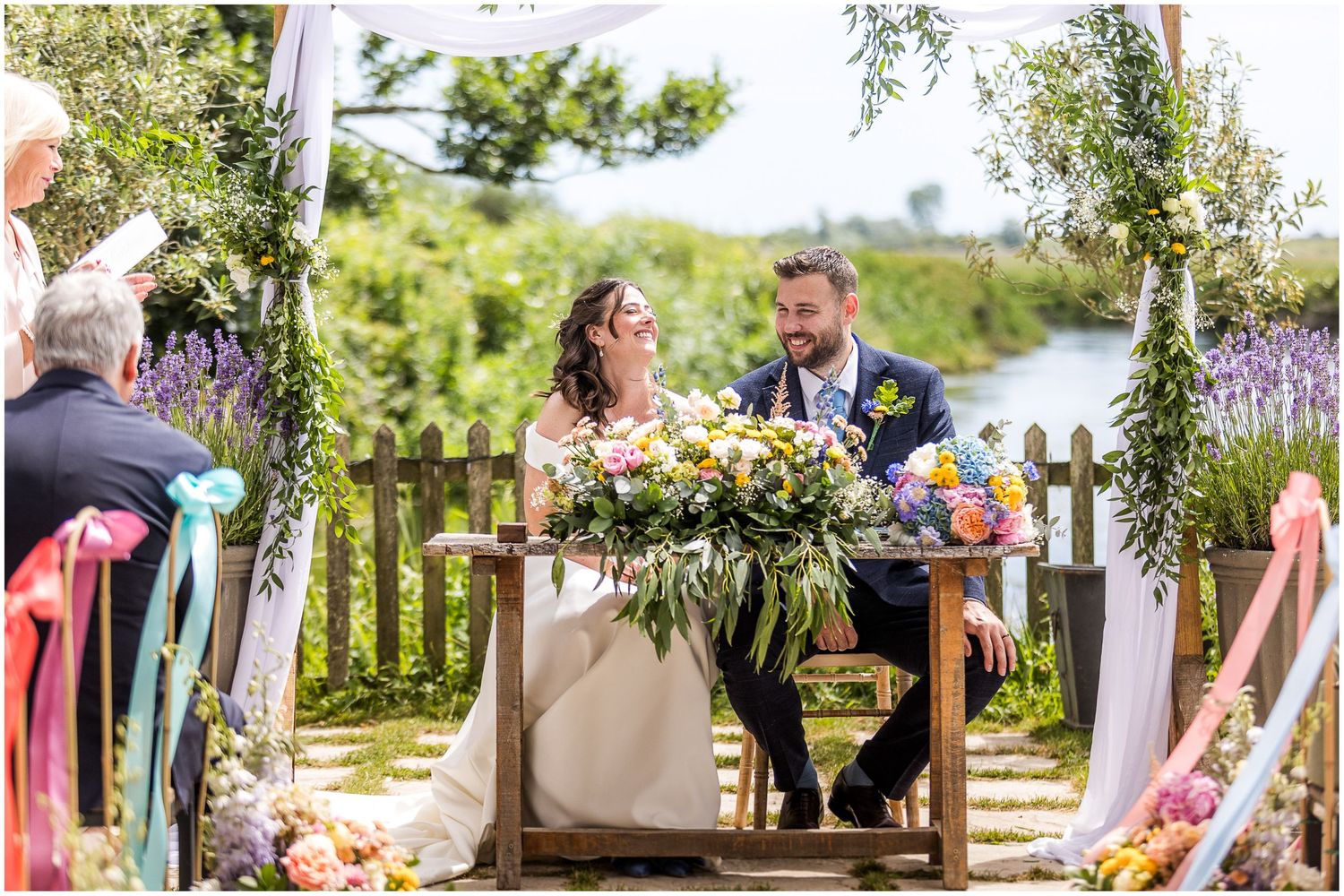 Bride and groom seated at outdoor wedding ceremony signing table decorated with colourful flowers and greenery and a countryside backdrop