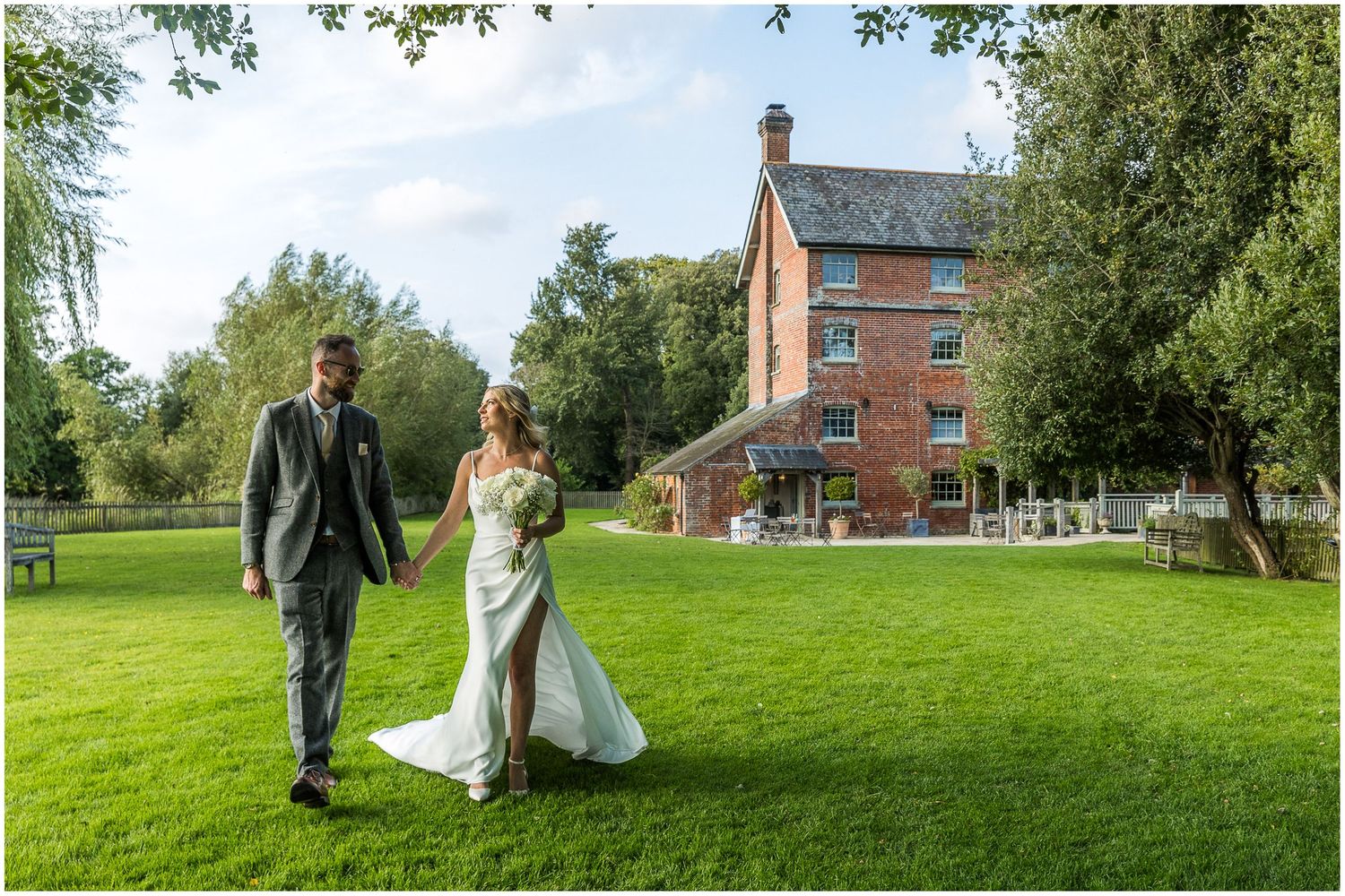 Panoramic wedding photography showing couple walking through scenic countryside with brick buildings.