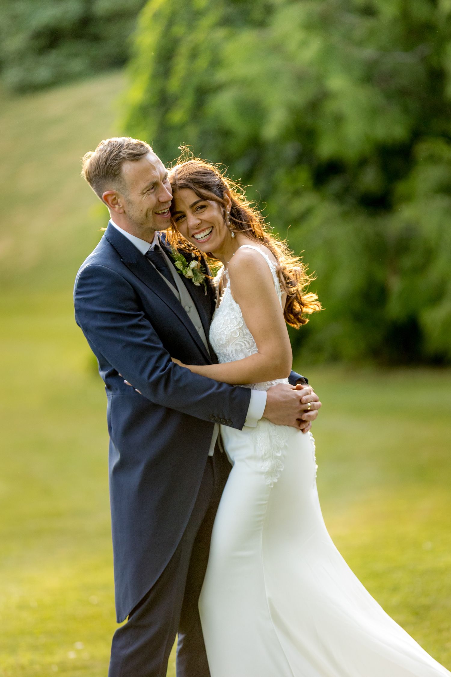 Wedding couple laughs together while posing on a grassy hillside.