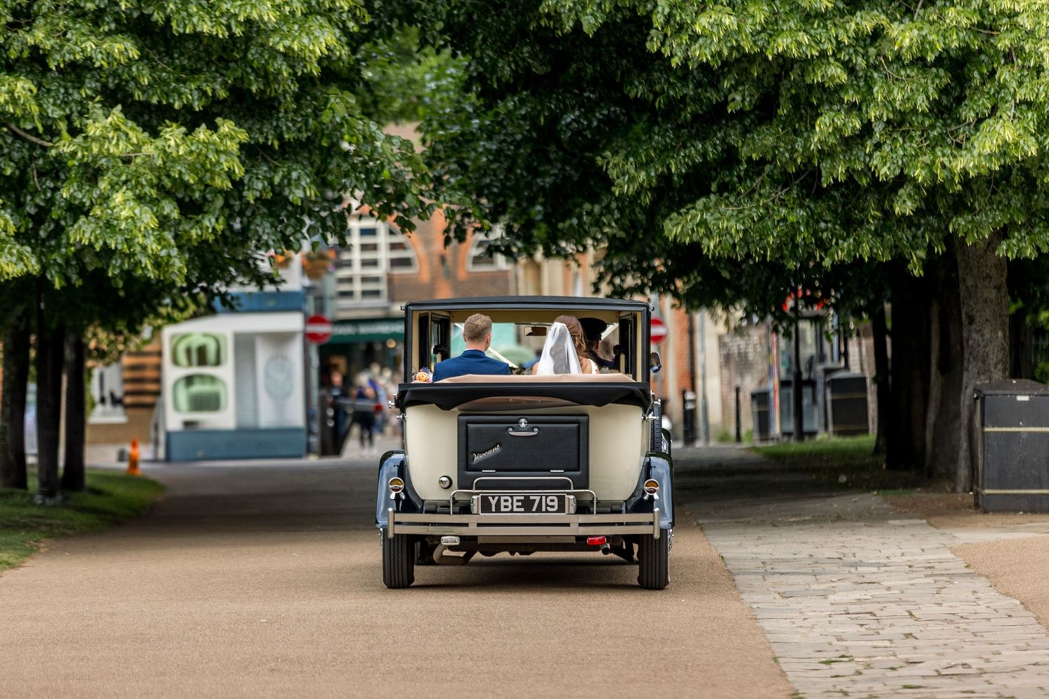Green trees frame wedding venue entrance with classic car visible in distance.