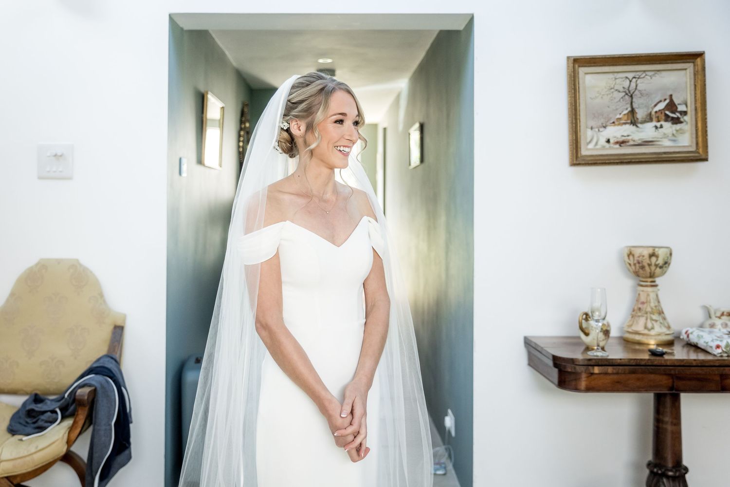 A bride in an elegant off-shoulder white gown and veil stands in a hallway.