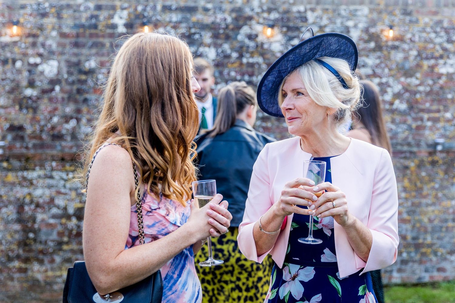 Wedding attendees chat and hold beverages at a brick-walled venue during sunset.