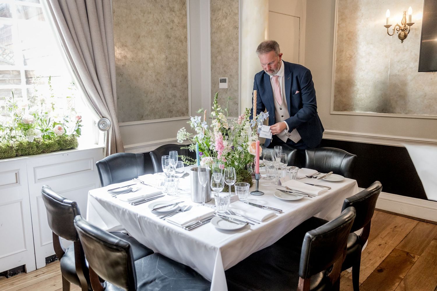 Elegant dining room setup with white tablecloths and place settings in a bright room with wallpaper.