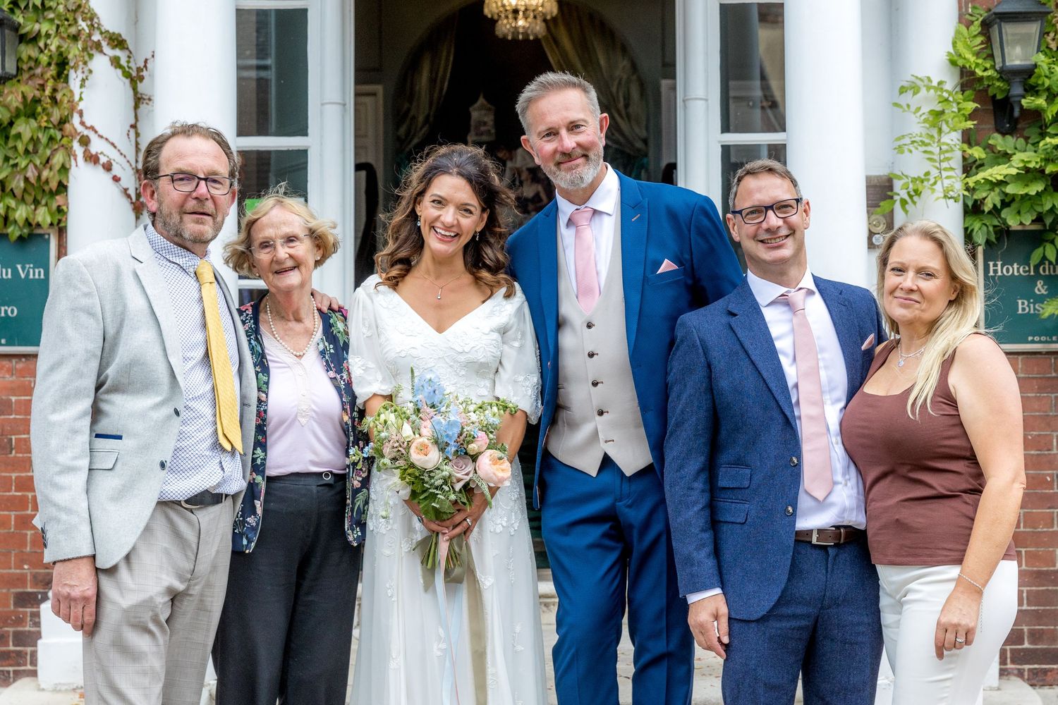 Wedding group photo outside an elegant brick venue with white pillars and greenery.