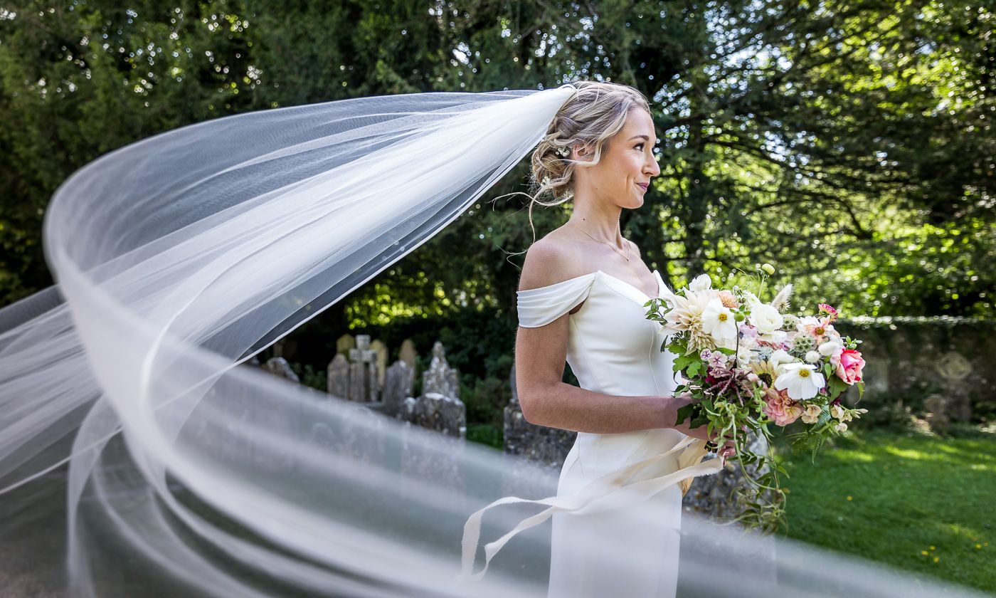 Flowing white wedding veil catches breeze in sunlit garden setting.