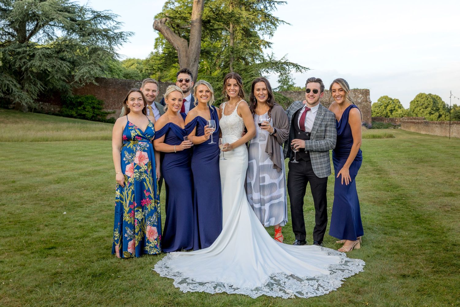 Wedding party poses for group photos on expansive lawn near historic stone building.