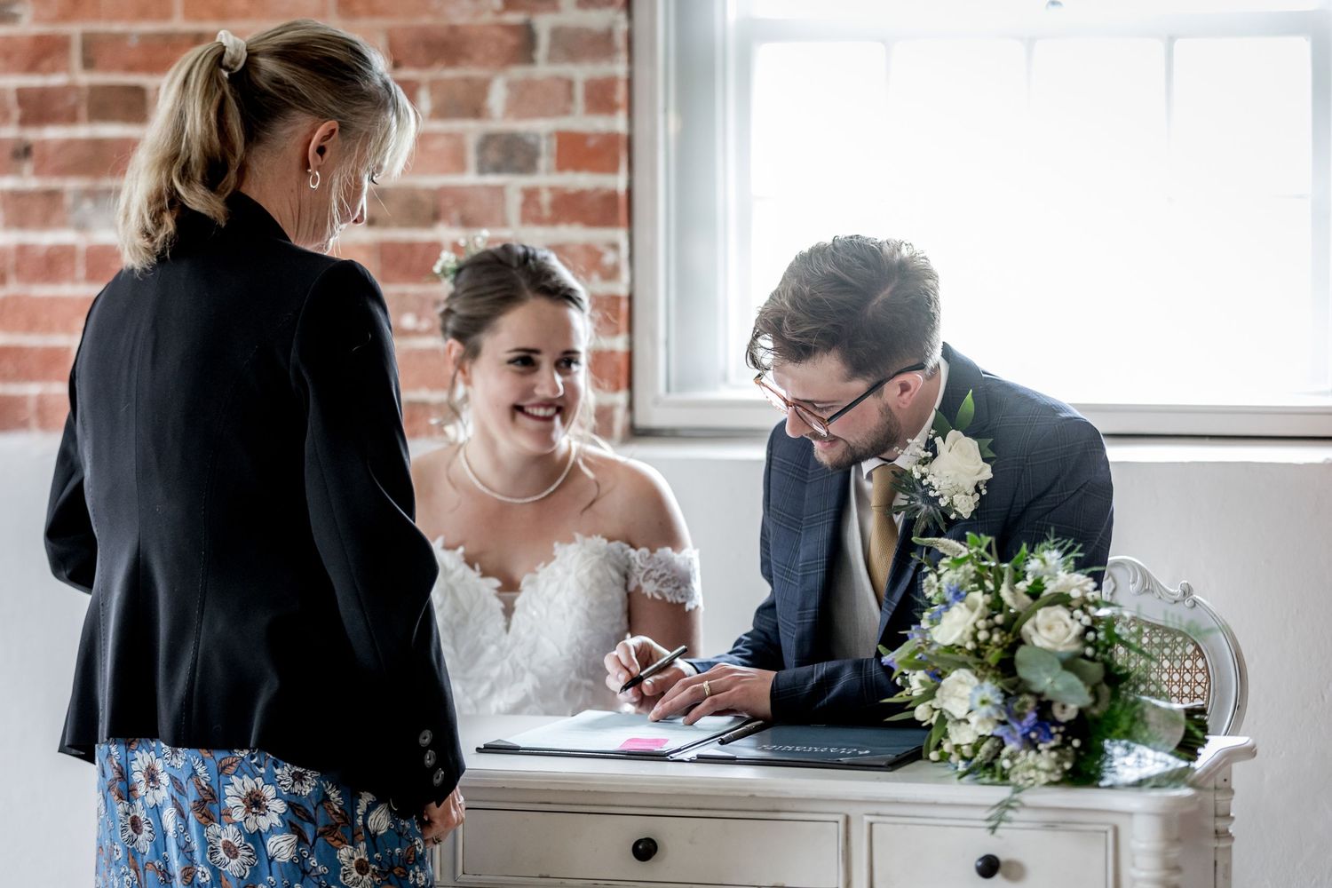Wedding ceremony in a rustic brick-walled venue with guests watching the couple sign documents.
