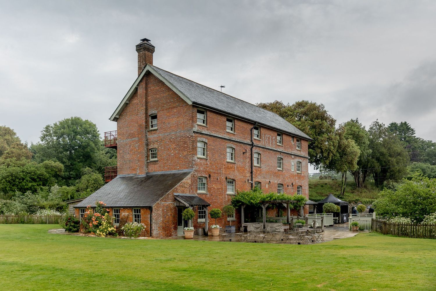 Row of rustic brick buildings and barns set against green countryside landscape.