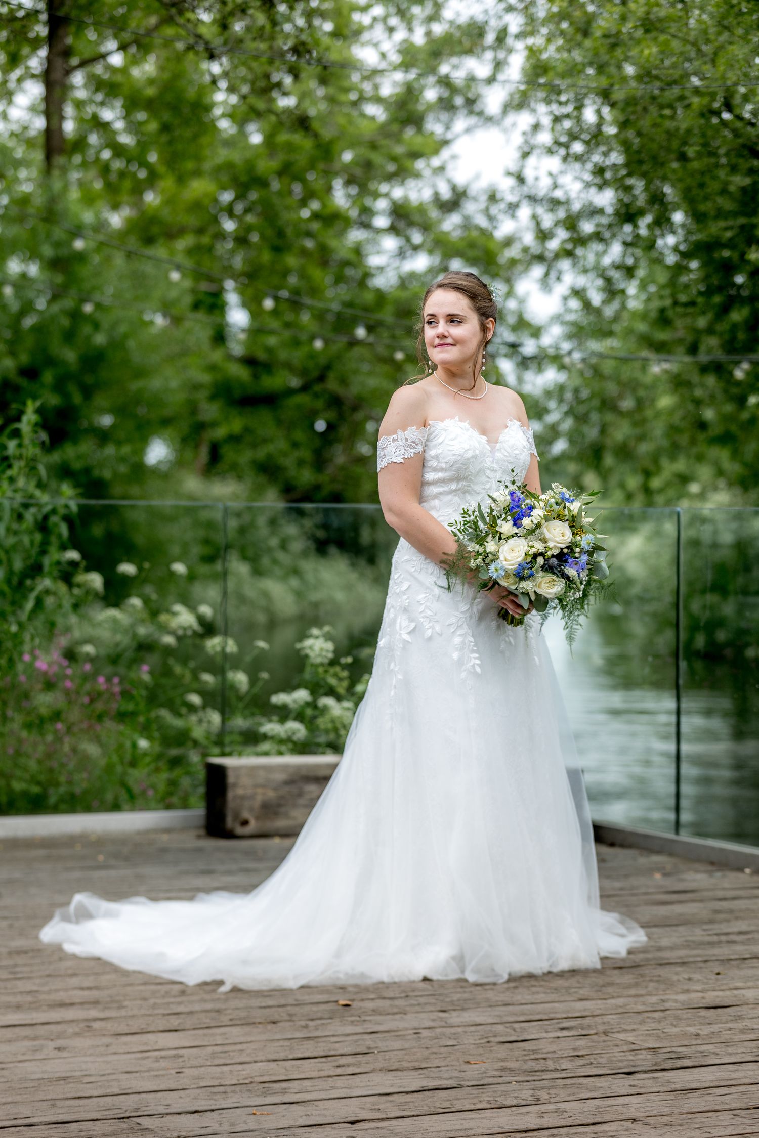 Bride in flowing white gown holds a vibrant blue and white bouquet on wooden deck.