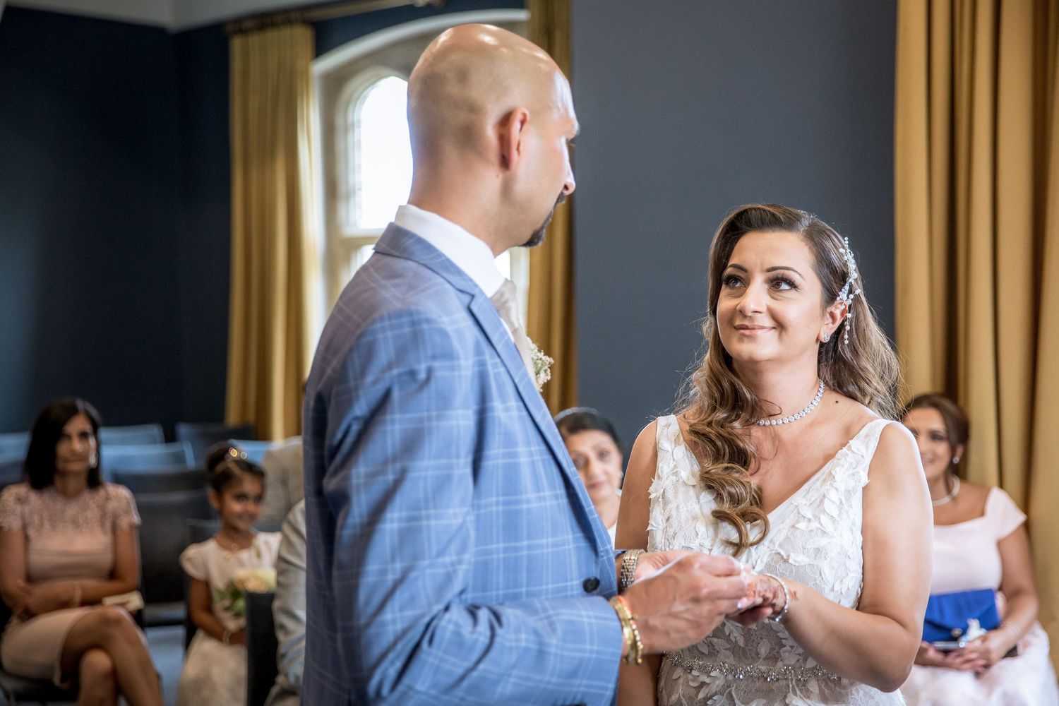 A couple shares their first dance as newlyweds in a sunlit ballroom with guests watching from the sides.