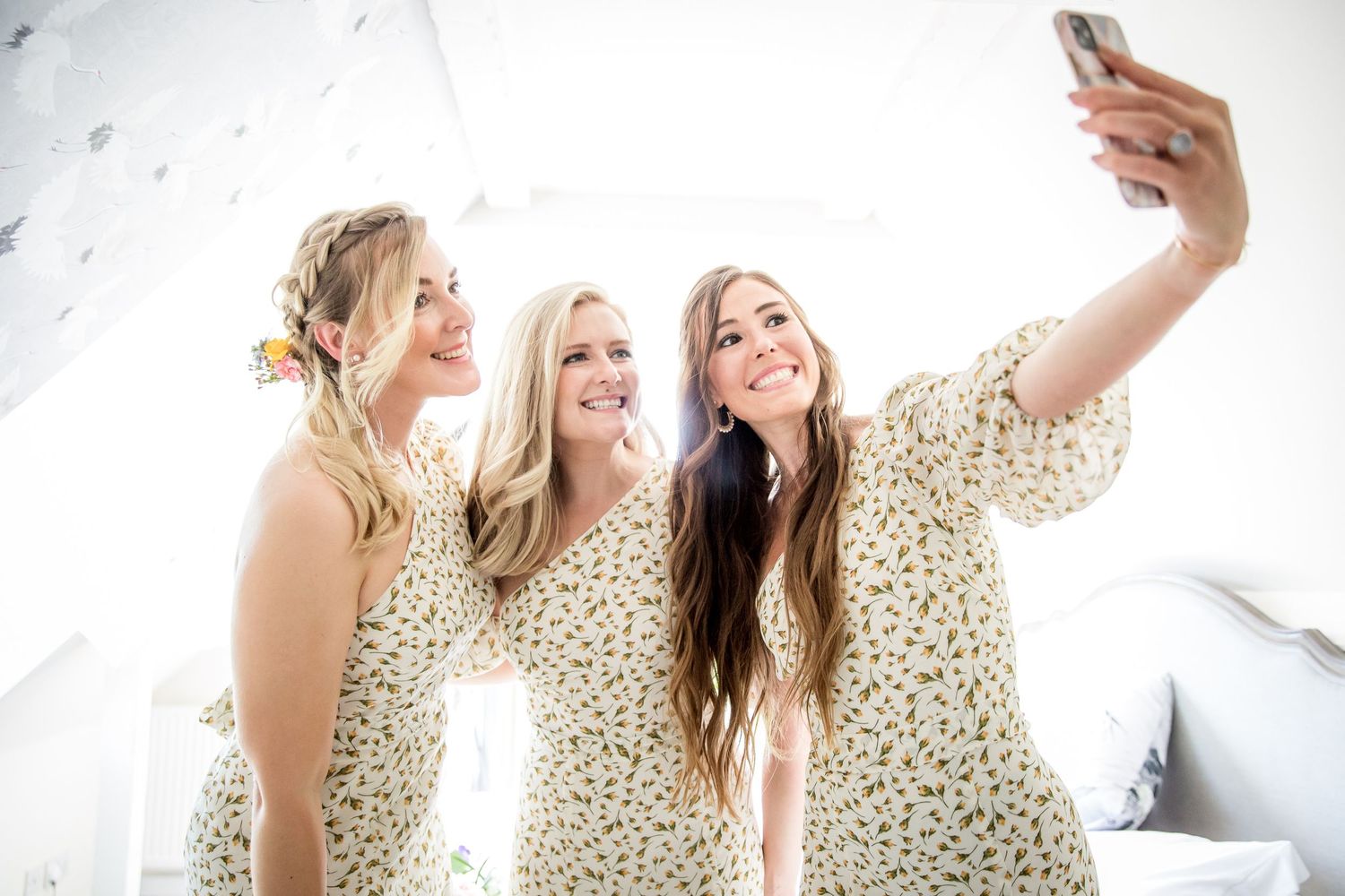 Three people wearing matching floral dresses take a selfie together while smiling and posing on a light background.