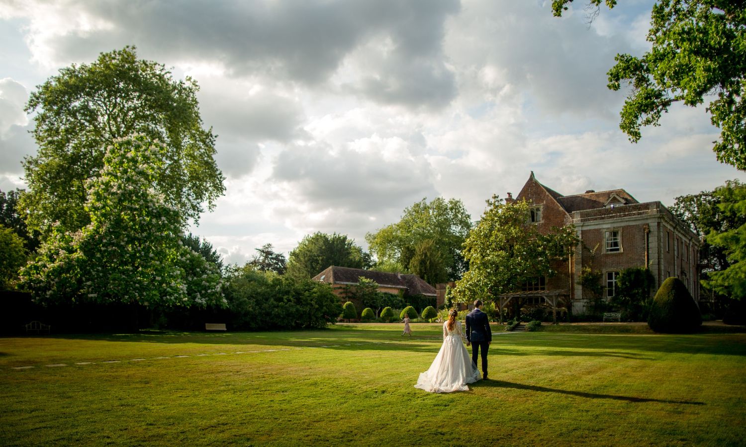 Couple stands on expansive lawn with historic mansion and cloudy sky in background.