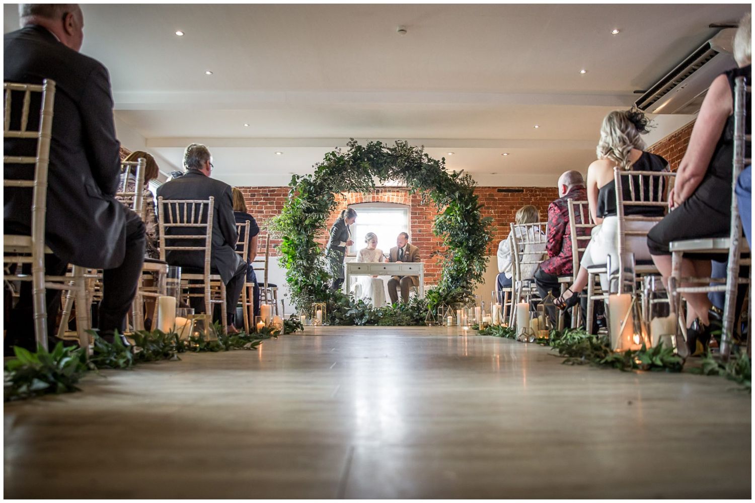Wedding ceremony aisle view with floral arch and guests seated in elegant chairs at an indoor venue.