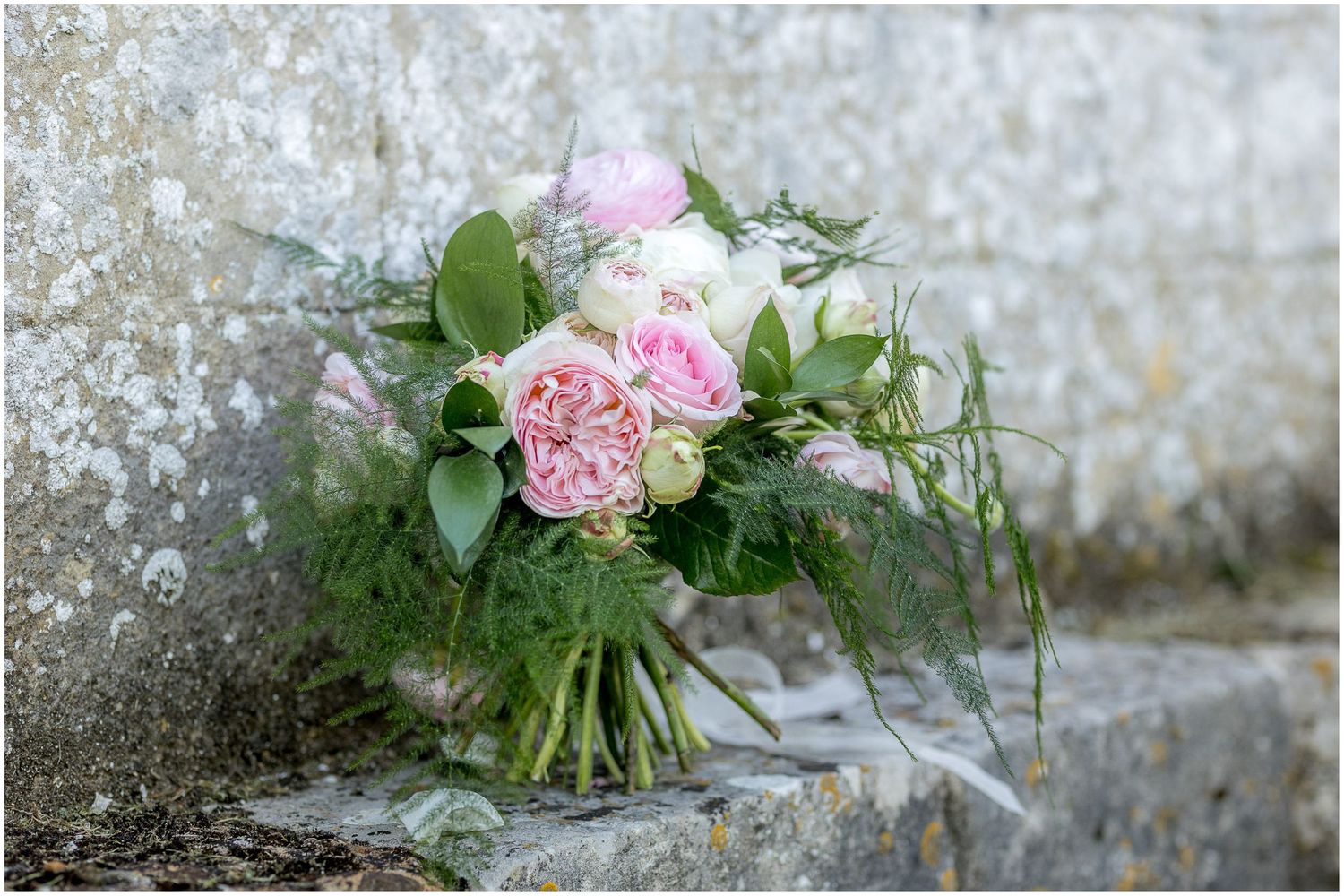 Pink floral wedding bouquet with roses and greenery resting on weathered stone ledge.