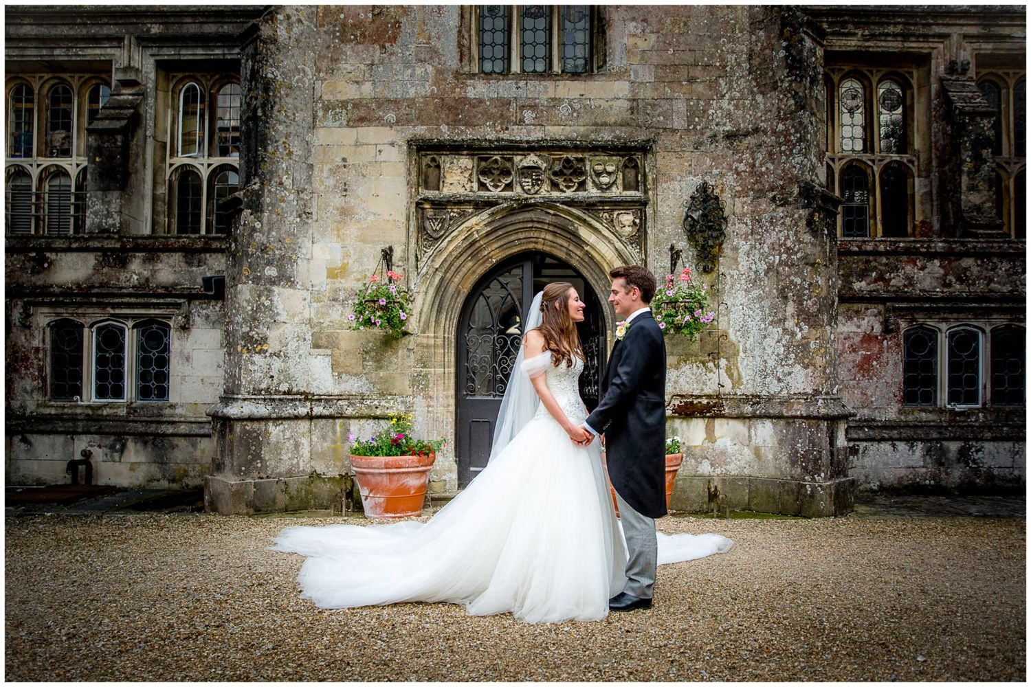 A couple in wedding attire stands in front of an ornate stone archway at a historic castle or estate.