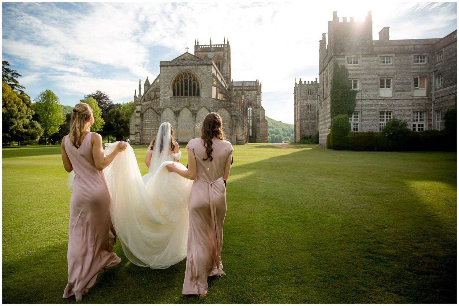 Wedding party in pink dresses lifts bride's dress on sunny lawn with elegant stone mansion in background.
