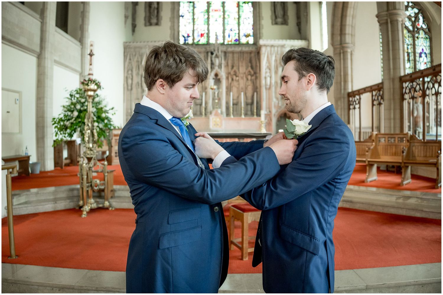 Two people in navy suits share an intimate moment in a sunlit church with stained glass windows.