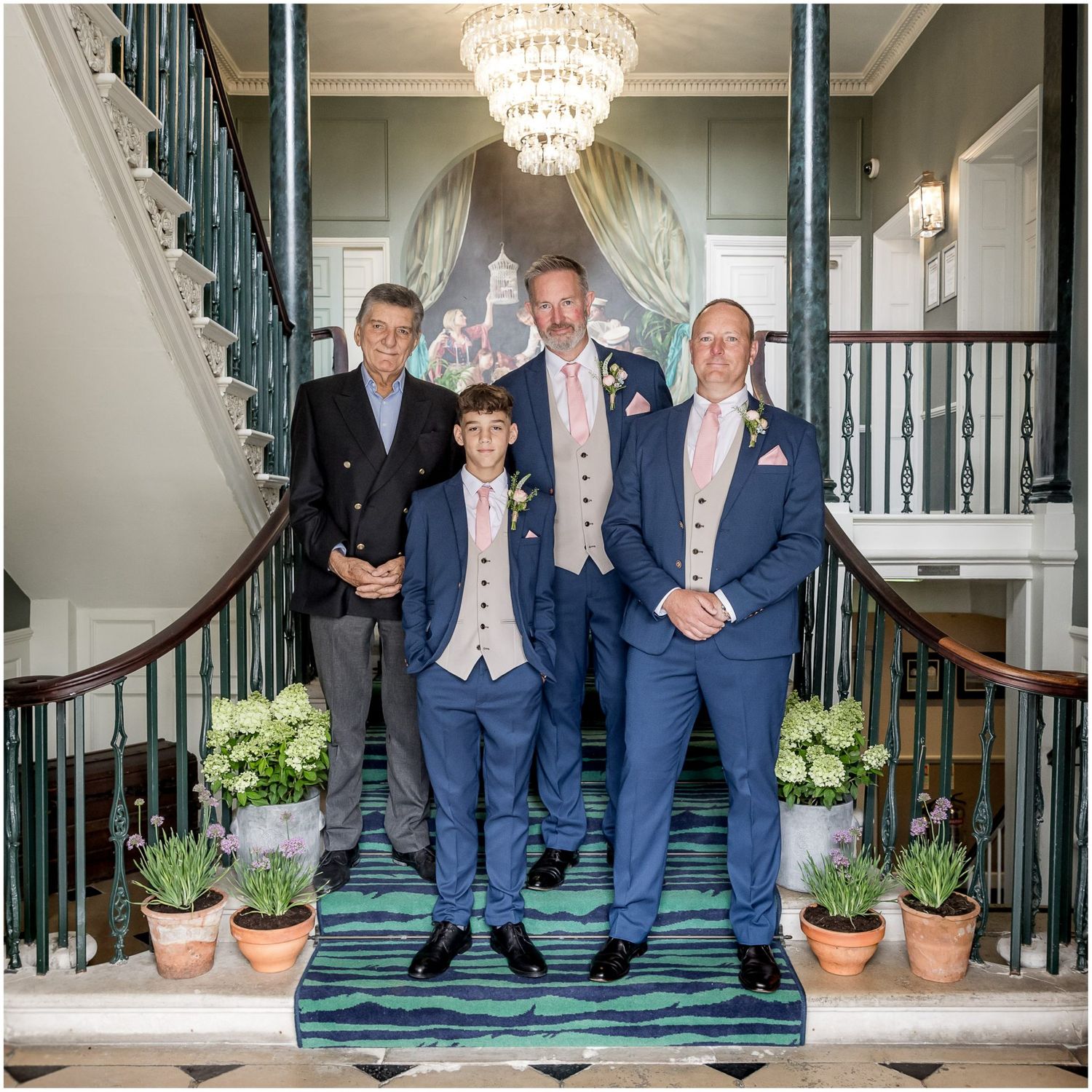 Wedding party members in navy suits pose on grand staircase with crystal chandelier.