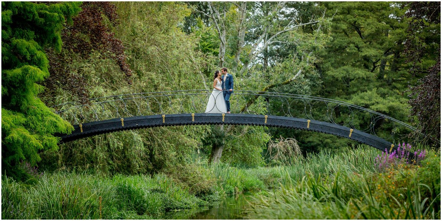 Artistic wide shot of couple standing on ornate garden bridge surrounded by greenery.