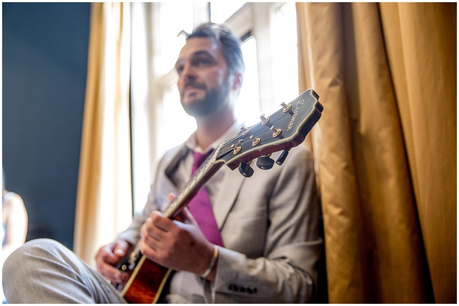 Musician in beige suit plays acoustic guitar by window with soft natural lighting and brown curtains in background.