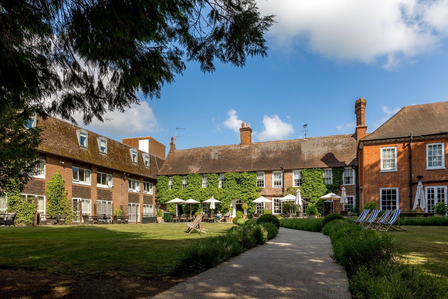 Traditional English manor house with manicured courtyard garden and brick buildings under cloudy sky.