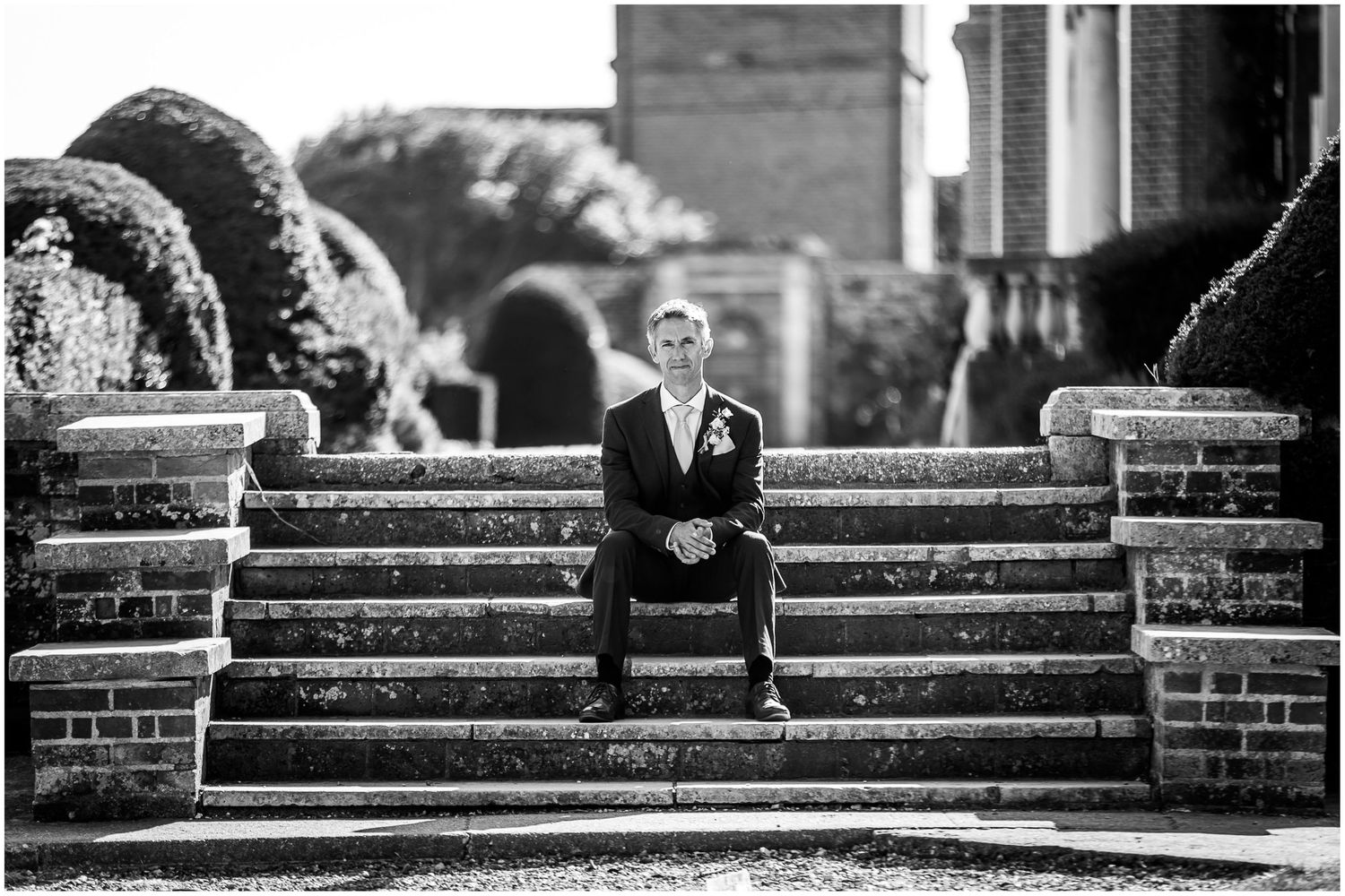 Black and white wedding photo of a groom sat on stone steps in the grounds of Walhampton School