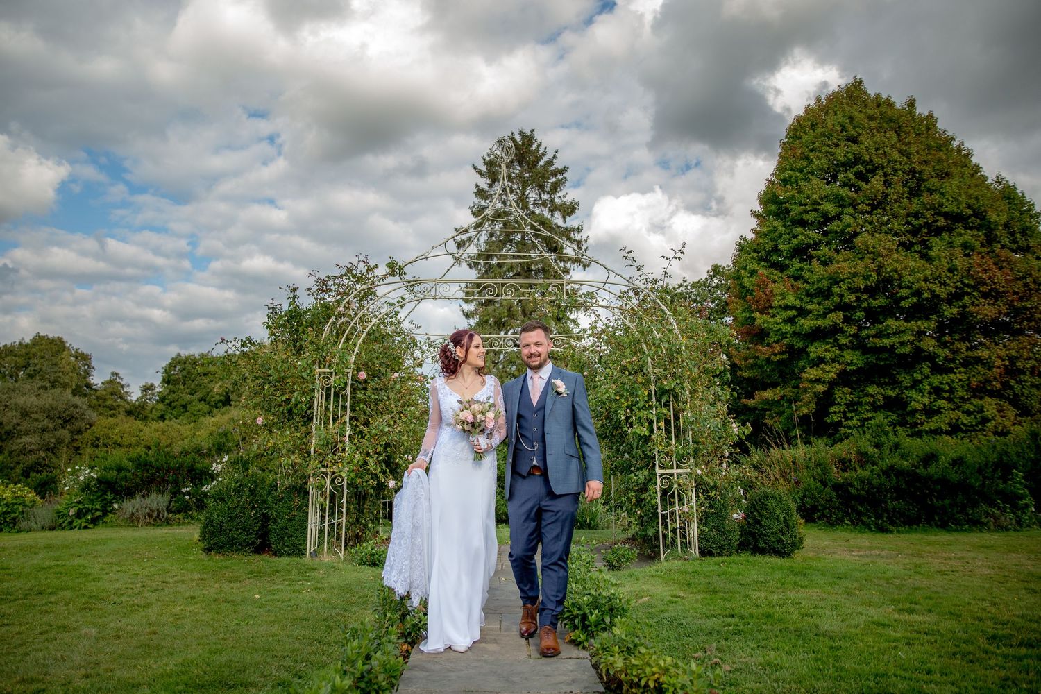 Wedding couple poses in a lush garden setting with dramatic cloudy sky overhead.