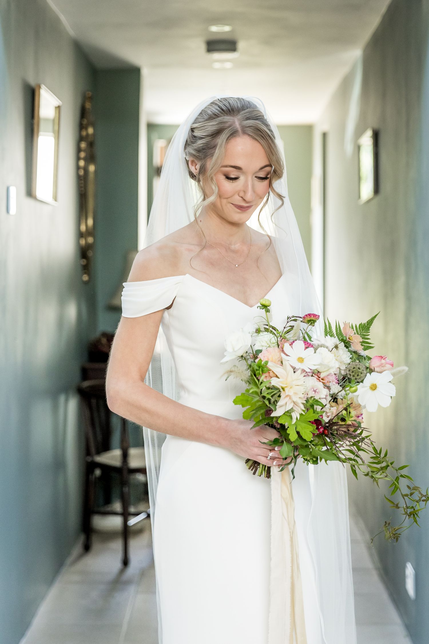A bride in an off-shoulder gown holds a delicate bouquet while looking down.