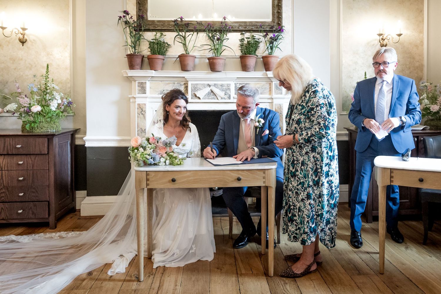 Wedding guests gather around a signing table in an elegant room with a fireplace.