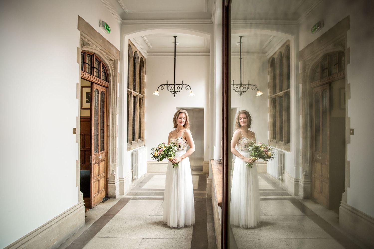Two people walk down an elegant white hallway with ornate architectural details and natural light streaming in.