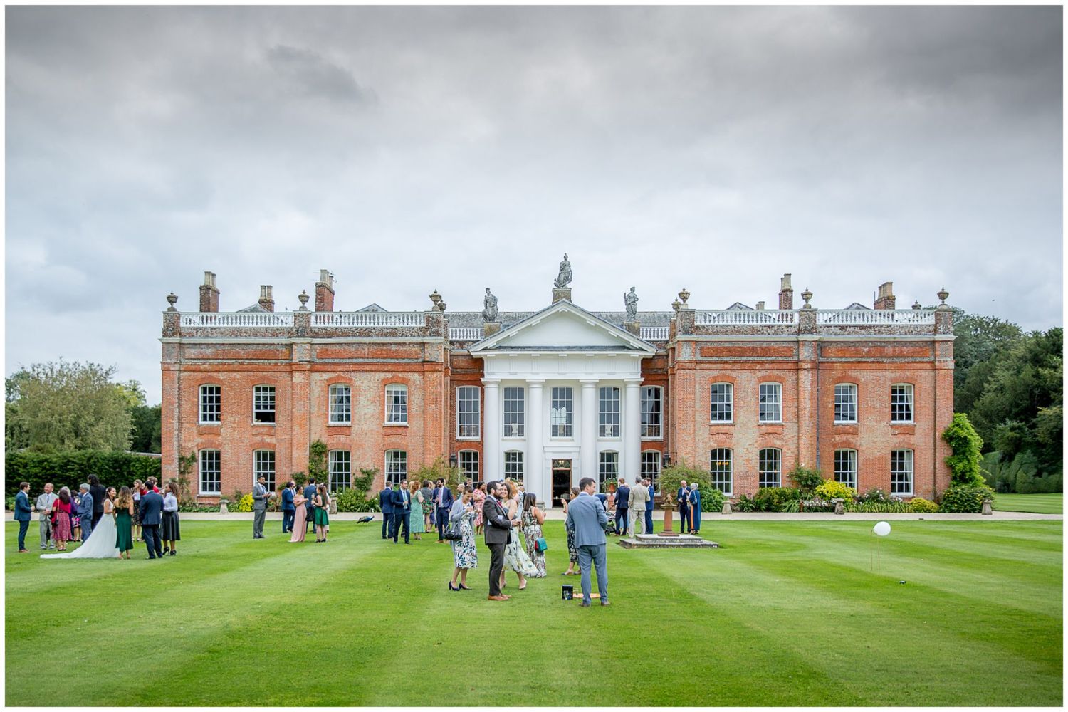 Panoramic view of an elegant red brick Georgian mansion with guests gathered on the expansive lawn.