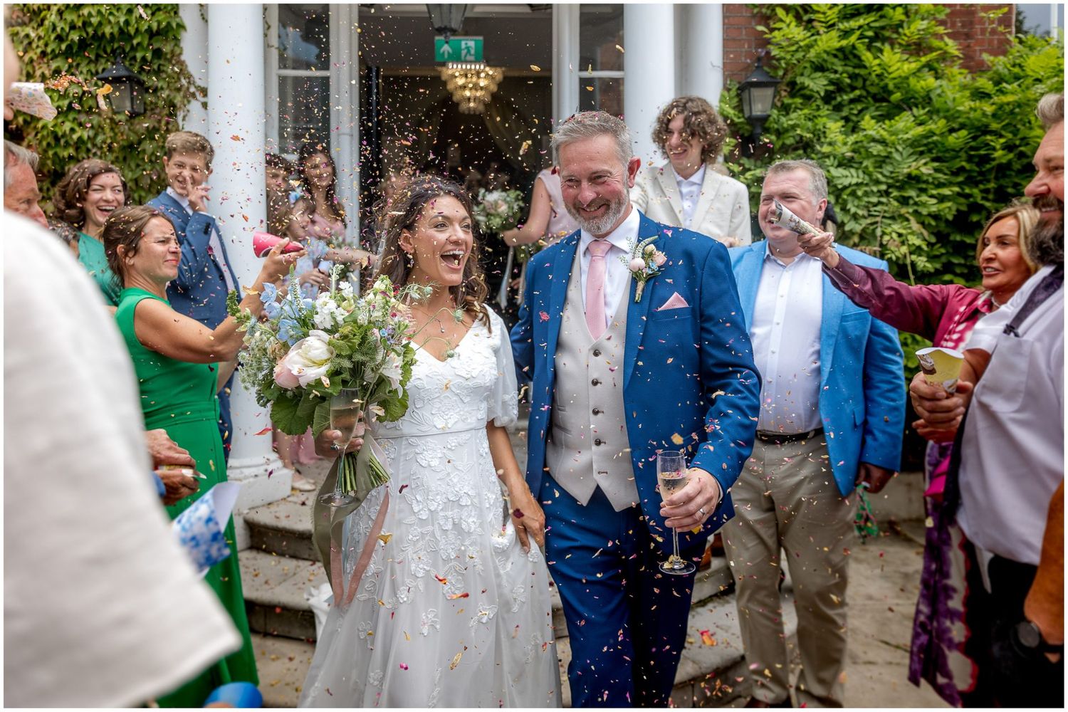 Wedding celebration outside a white columned building with guests throwing confetti.