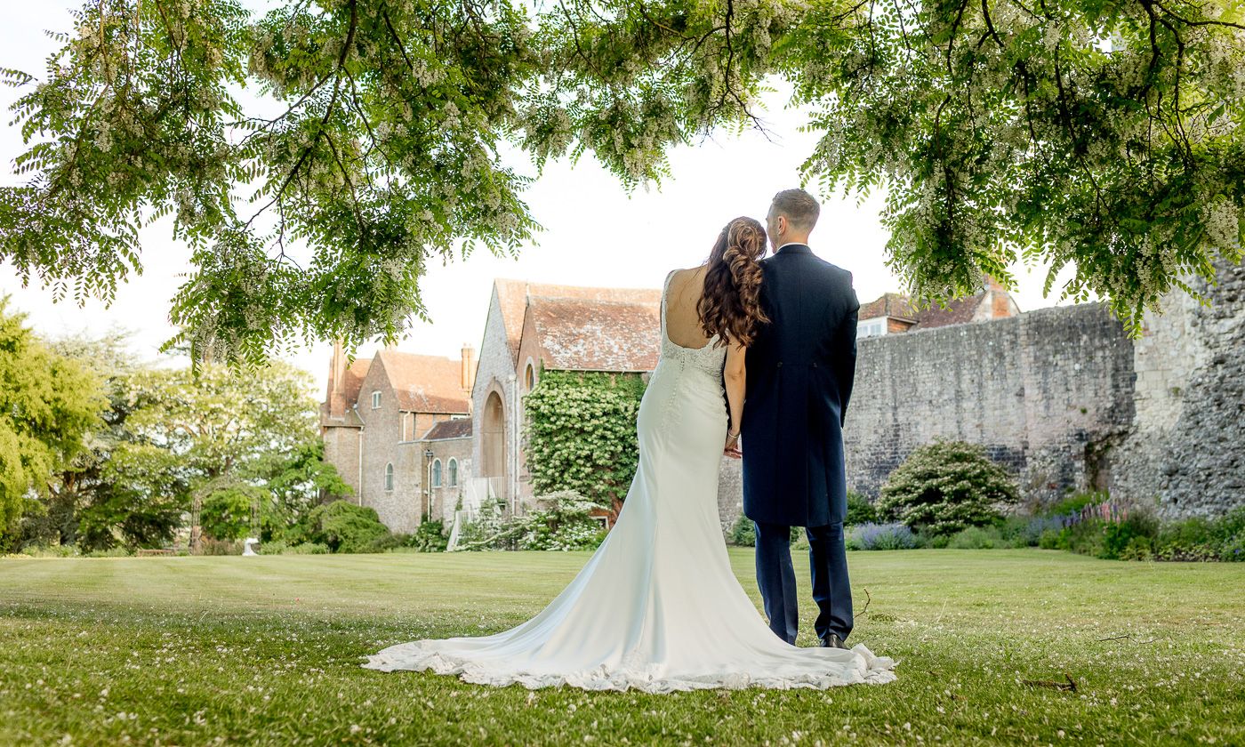 Wedding couple walking through lush garden setting with historic building in background.