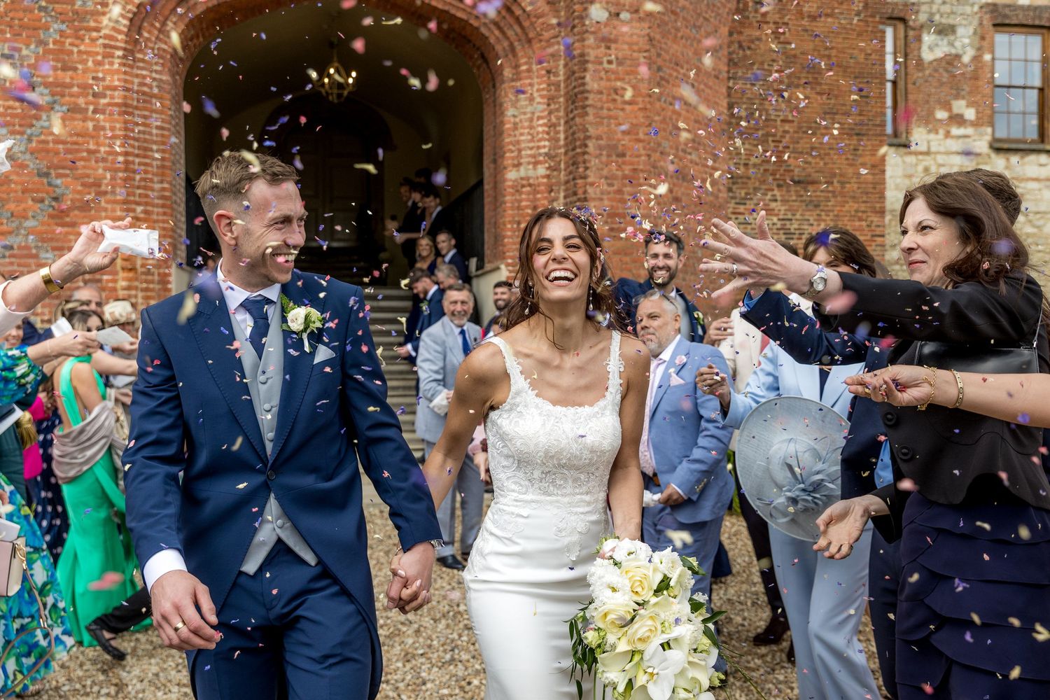 Guests throw confetti at a wedding celebration outside a rustic brick building.