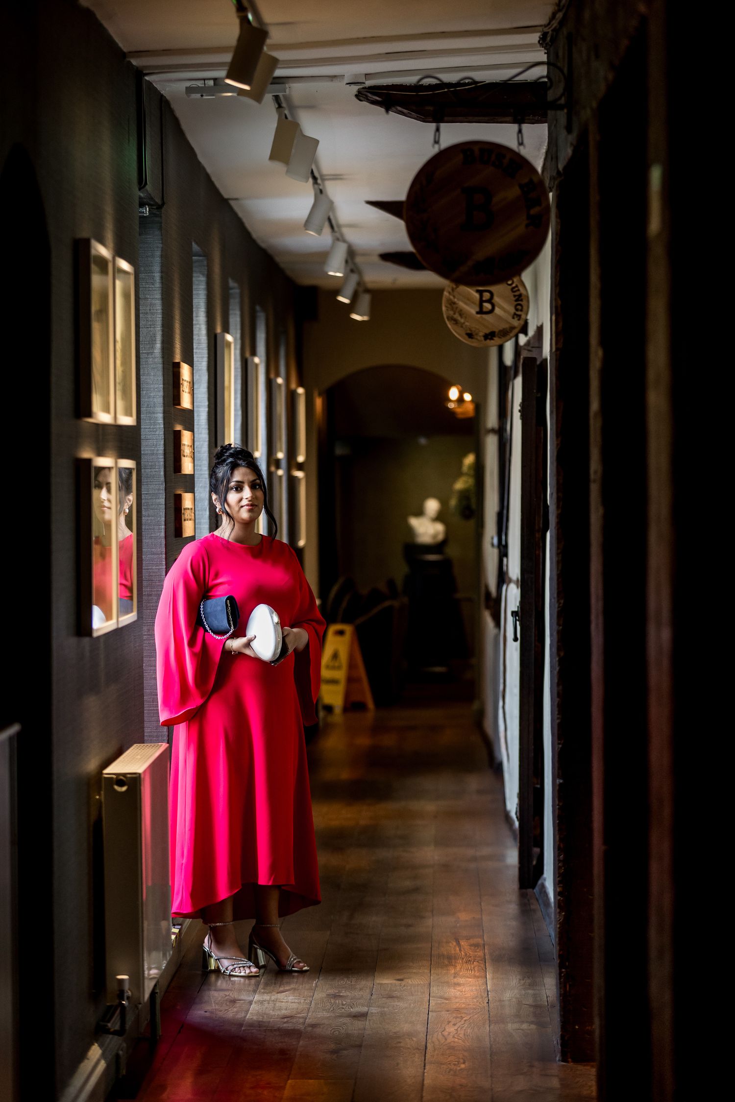 Woman in flowing red dress poses dramatically in a long dark hallway with vintage lighting.