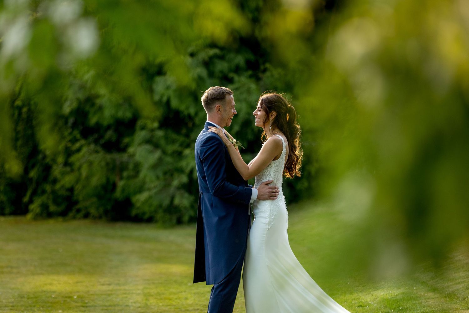 Romantic wedding couple embraces on a lush green lawn with trees in the background.